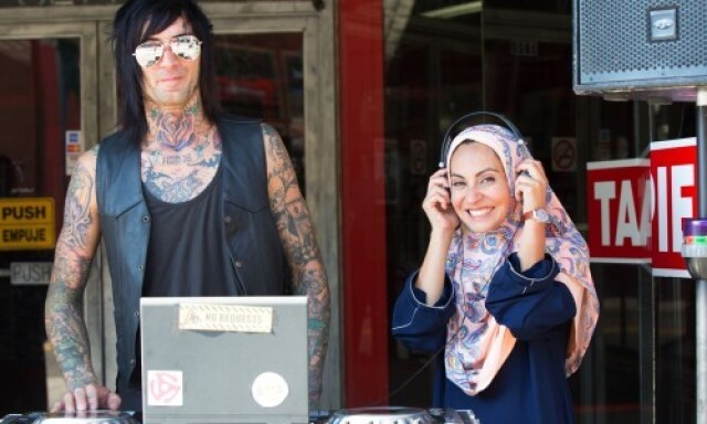 A man covered in tattoos standing next to Muslim woman wearing a hijab and headphones. They are smiling next to a DJ turntable.