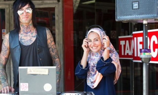 A man covered in tattoos standing next to Muslim woman wearing a hijab and headphones. They are smiling next to a DJ turntable.