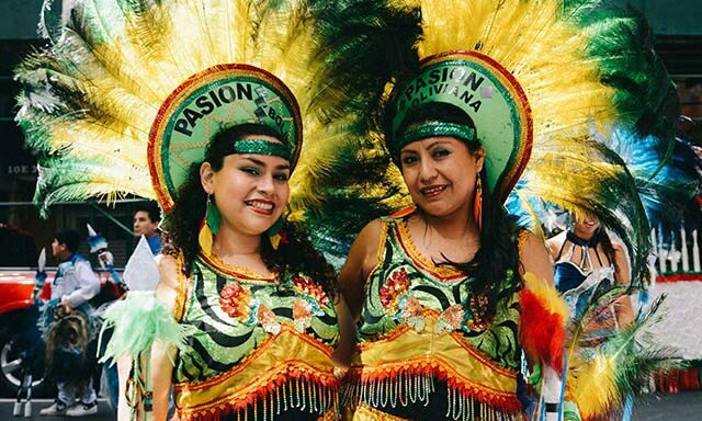 Two smiling Bolivian women wearing elaborate green, yellow, and red costume dresses and feathered headdresses that say Pasion Boliviana.